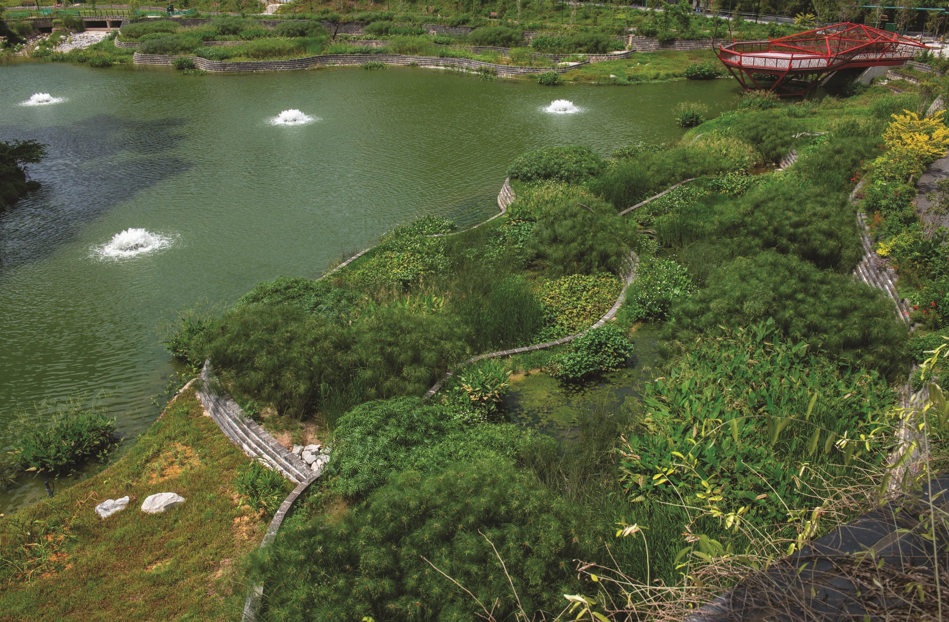 Aerial view of a green pond with fountains, landscaped banks, and a red viewing platform in a park.